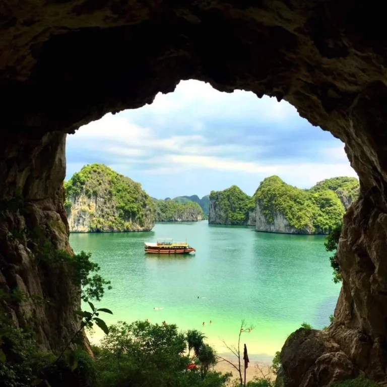 View of emerald green waters in Ha Long Bay, framed by the mouth of a limestone cave, with towering karst mountains and a traditional boat floating in the background.